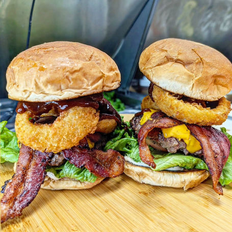 Two ridiculously overstuffed bacon cheeseburgers with huge onion rings in the stacks, side by side on a cutting board, lots of lettuce and sauce, bacon hanging out like big long tongues, both twice as tall as they are wide.