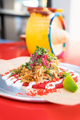 Side view of a very colorful mini-tower of shredded ingredients garnished with pickled radish and cilantro and lime wedge, con crema on a soft tortilla, laying on a metal plate on a red tablecloth, blurry Mexican hat wheel visible in the background.