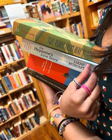 The by-now-familiar woman in the blue polka-dot dress, but in extreme closeup of her hands and wrists only, covered in costume jewelry, spine labels of the books clearly visible: Two Roads From Here, The Hypnotist's Love Story, Ducks Newburyport by Lucy Ellmore, and Miss Peregrine's Home for Peculiar...