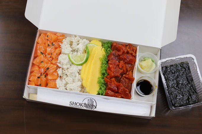 Straight-down view of and open white heavy-paper bento box filled with four sets of food: raw salmon chunks, shredded white something, maybe coconut, slices of mango, and deep red beef chunks; small cups of fish sauce and wasabi/ginger in the last cell, plus a Shokunin-branded chopstick package. And finally, a small clear-plastic container of dried seaweed just beside on a dark wood tabletop.