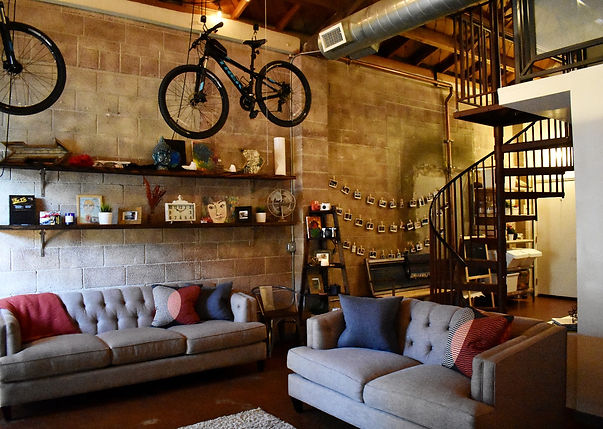 an interior view of a typical live-work space in Pomona CA, with a block side wall and a bike hanging from the ceiling, two couches and decorative shelving along the wall, exterior ducting visible above and open wood-rafter ceiling