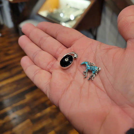 a hand holding out two pieces of earring jewelry in its palm, almost filling the frame, wooden flooring blurred below; looks like a topaz horse and black obsidian wheelio