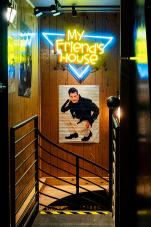 a glimpse of the downward spiral staircase leading down to My Friend's House speakeasy gaming bar below Rom with neon sign and poster and railing and woodgrain