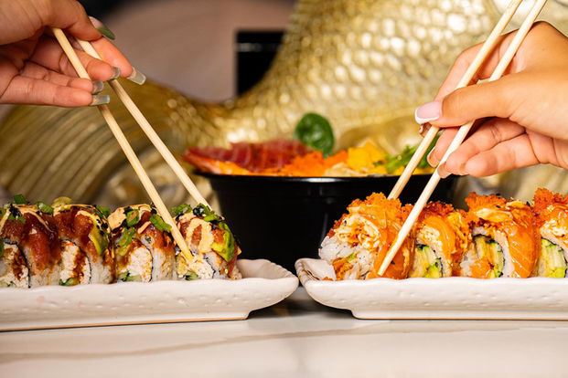 Closeup sideview of two women’s hands with chopsticks, each picking up their first sushi rolls from their side-by-side tray plates filled with more rolls, one salmon, the other heavily drizzled crabmeat and garnishes. Part of golden-fish sculpture can be seen in the blurry background.