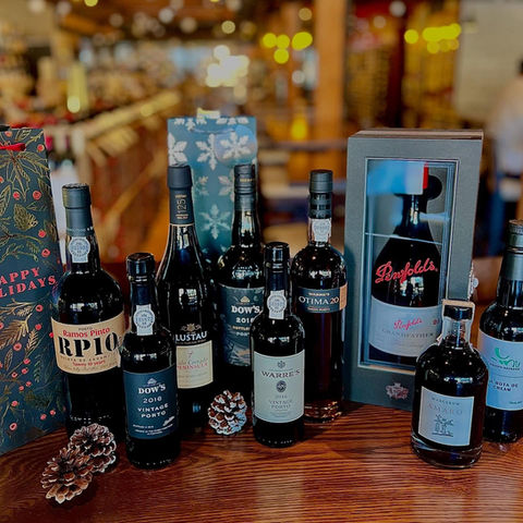 An array of gift bottles & boxes sitting atop a polished wooden surface, tightly packed together for display with some pinecones. Various bottle shapes & sizes, wines, liqueurs, brandies, etcetera.