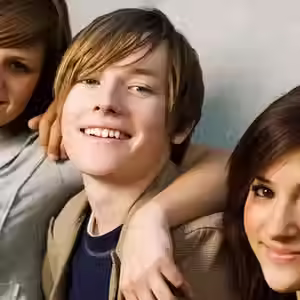 A group of young friends sits close together, smiling and enjoying each other's company against a concrete wall.