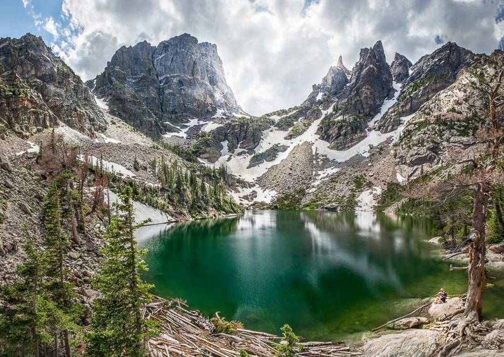 Green alpine lake below snowy jagged mountains, with pine trees and a lone hiker sitting on a rock under cloudy skies.