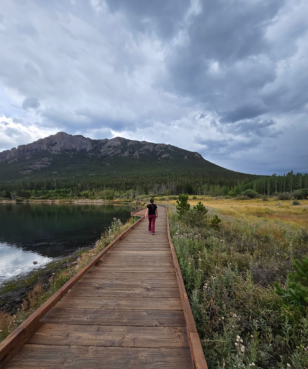 Person walking on a wooden boardwalk beside a calm lake, with mountains and dramatic cloudy sky in the background.