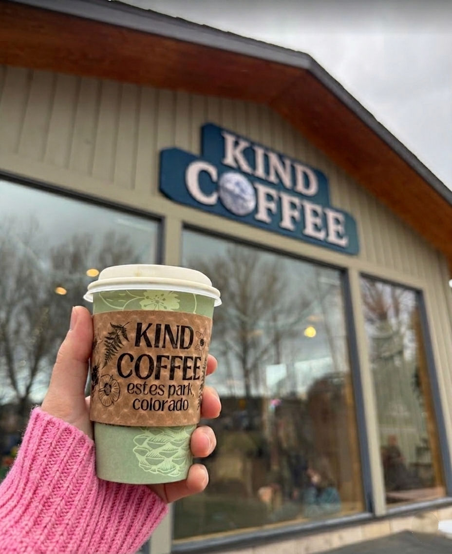A hand in pink sleeve holds a coffee cup labeled "Kind Coffee, Estes Park, Colorado" outside a shop with a "Kind Coffee" sign.
