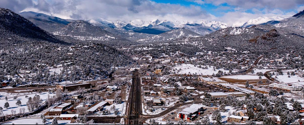 Aerial view of a snowy mountain town with a road running through the center, pine-covered hills, and snowcapped peaks.
