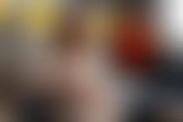 Mulher de olhos fechados se espreguiça numa sala de reunião, com pessoas desfocadas e parede cinza com formas amarelas.