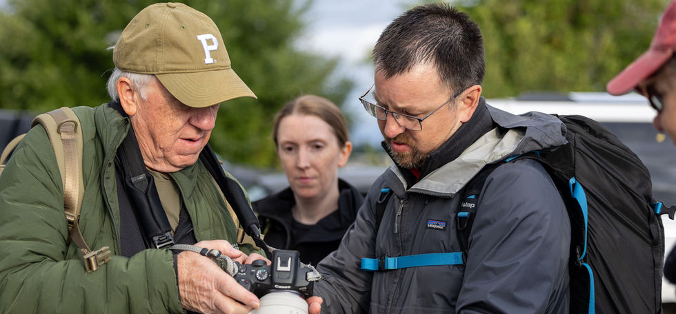 Two men examining a camera with lens outdoors, other people watching nearby.