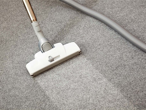 Vacuum cleaner on gray carpet showing a clean, light stripe. Background has a coiled hose. Mood is tidy and organized.