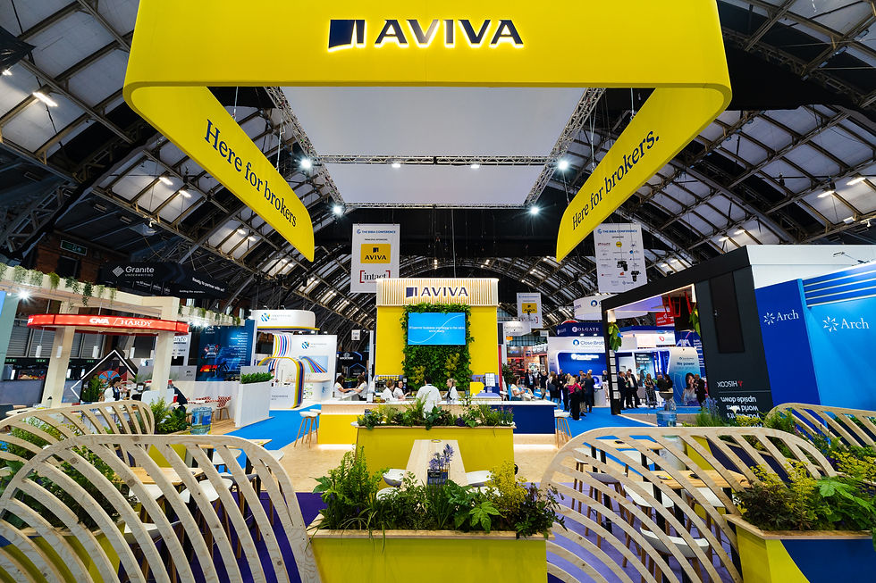Bright AVIVA exhibition stand in an exhibition hall, with yellow hanging banners reading 'Here for brokers'. Attendees milling around below.