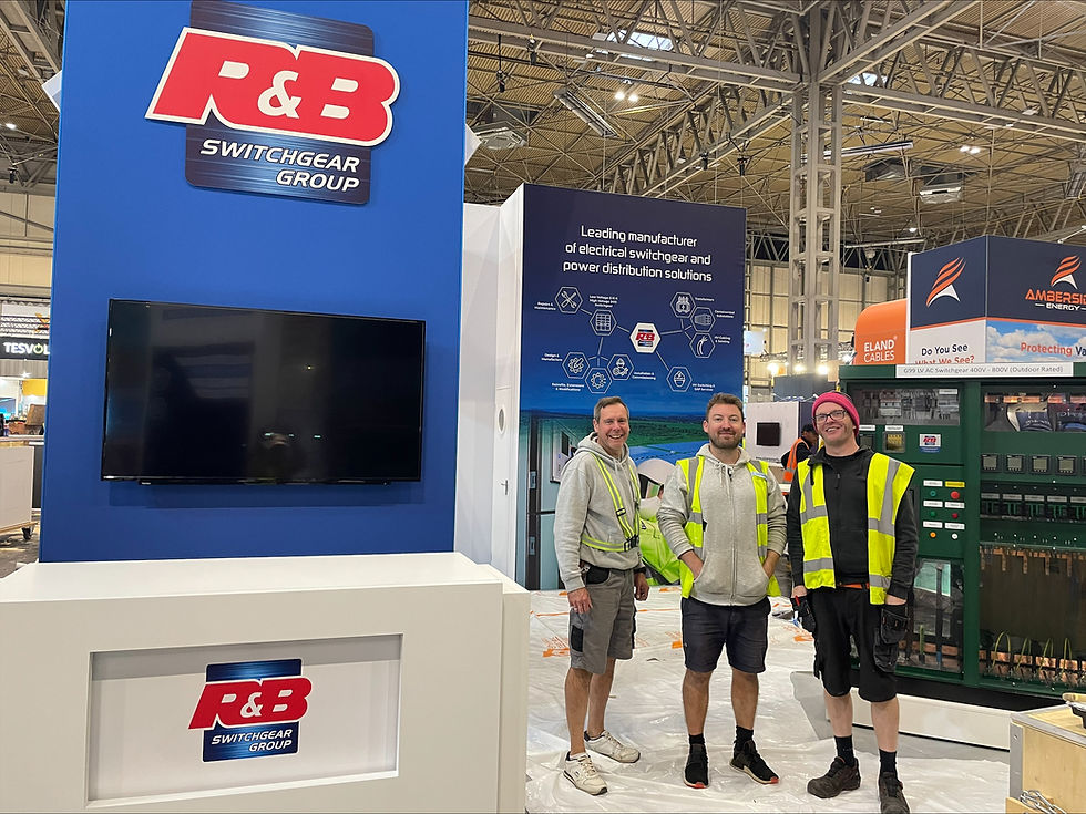 Three exhibition stand builders wearing hi vis jackets stand smiling on an exhibition stand for R&B Switchgear.