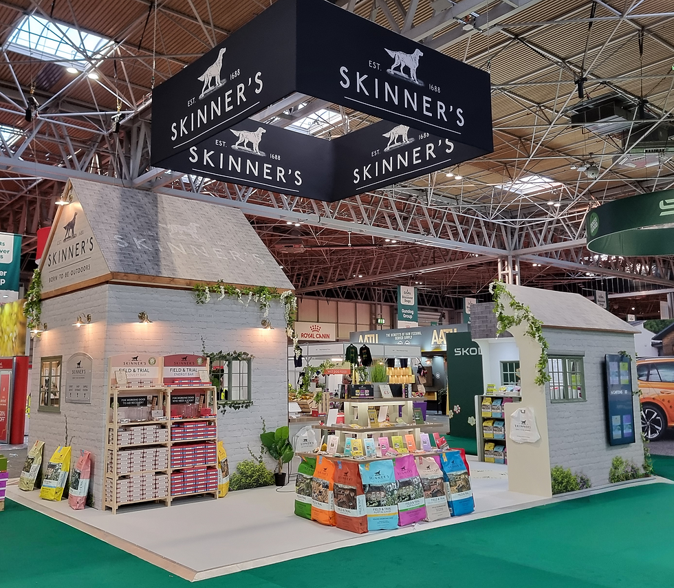 Skinner's impressive exhibition stand at Crufts. It is shaped like a cottage, with a shed outhouse, and an area for displaying dog food in the centre of the space. It has warm lighting and is laid out below a banner reading 'Skinner's'.