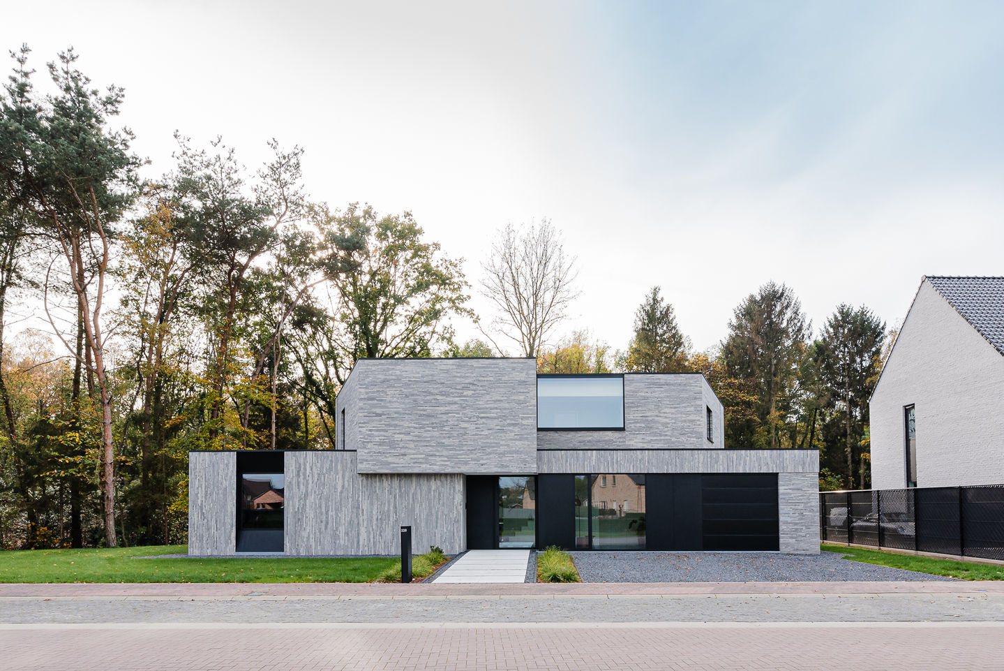 Moderne woning in Diepenbeek met een strakke, grijze gevelsteen van het type MBI Geostylistix shaded white. De woning heeft aluminium ramen en een plat dak met schuine muren, waarbij de ramen doorlopen tot aan de dakrand. De tuininrichting is harmonieus geïntegreerd in de omliggende bosrijke omgeving, wat bijdraagt aan een serene en eigentijdse uitstraling