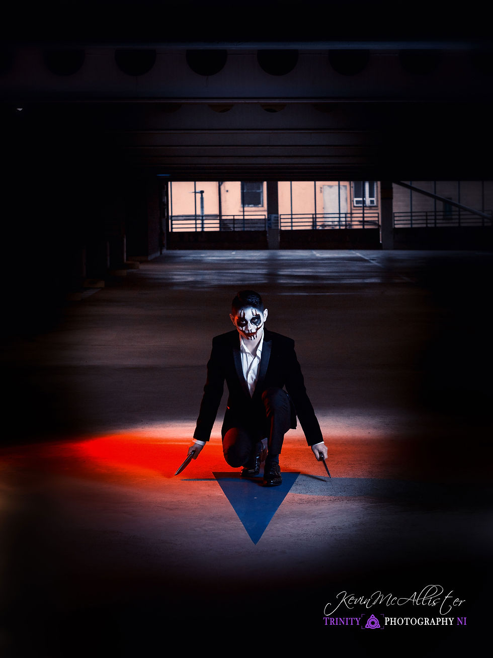 A person in a suit and face paint kneels in a dim garage, holding two knives. Red light casts dramatic shadows, creating an eerie atmosphere.