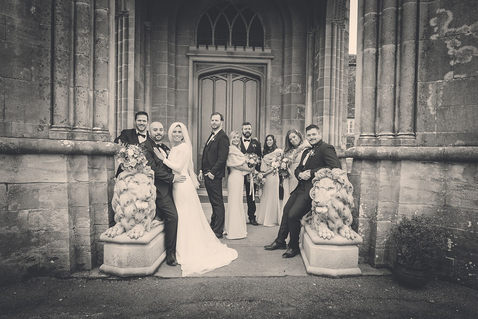 Bride, groom, and wedding party pose by stone lions outside a large stone building. Everyone is smiling and holding flowers. Black and white photo.