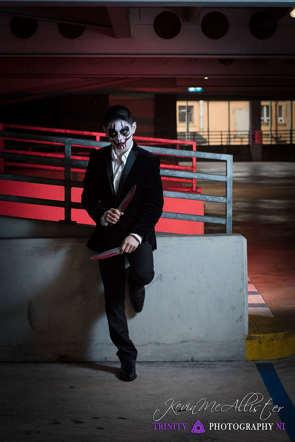 Person with scary makeup in a suit holds knives, leaning against a wall in a dimly lit parking garage with red accents. Eerie mood.