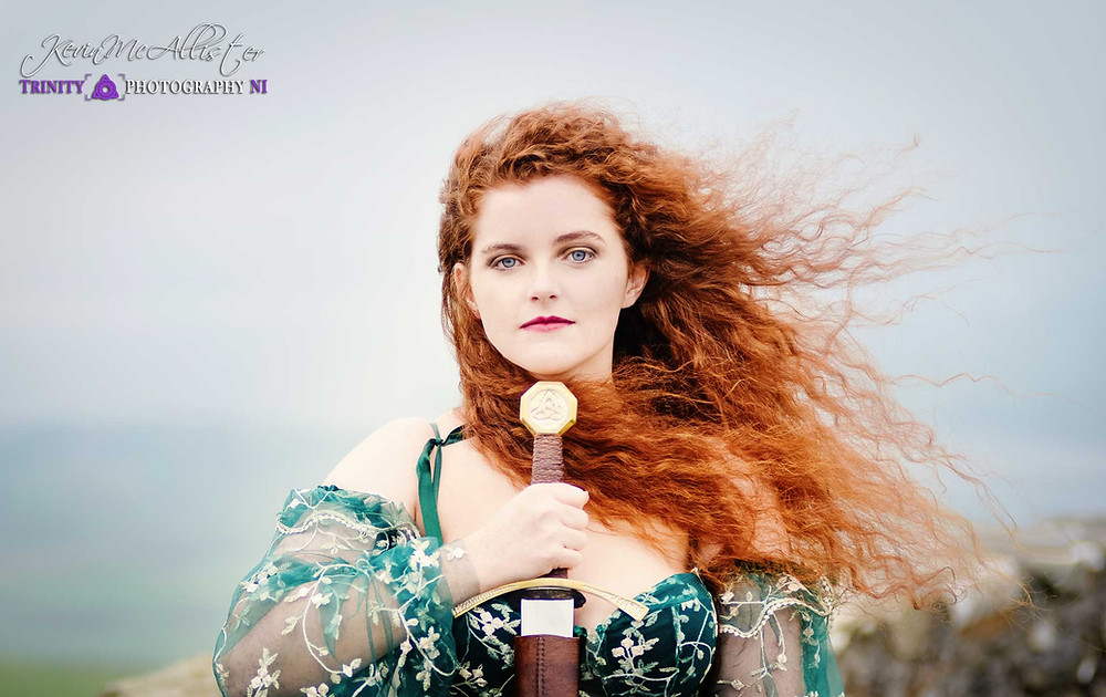 Redheaded model in green dress holding a sword whilst standing on a mountain top