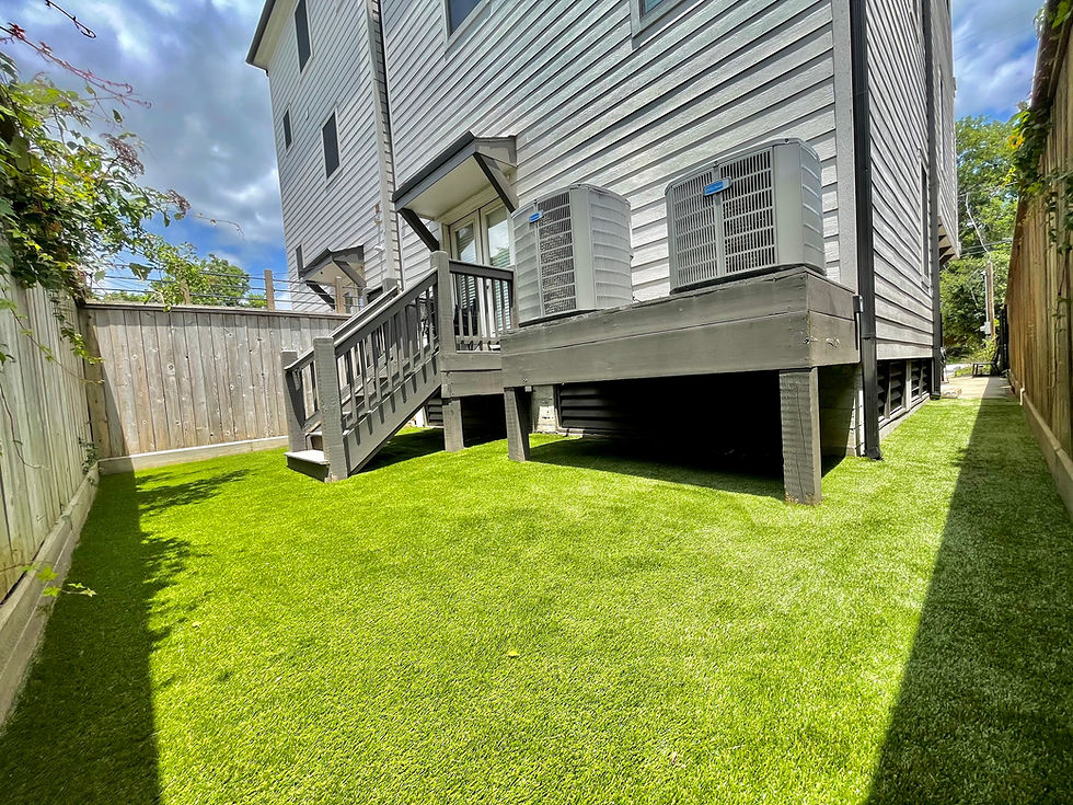 Backyard with lush green grass, a wooden deck, air conditioning units, and a grey house exterior. Fence and greenery on a sunny day. Downsides of Artificial Turf