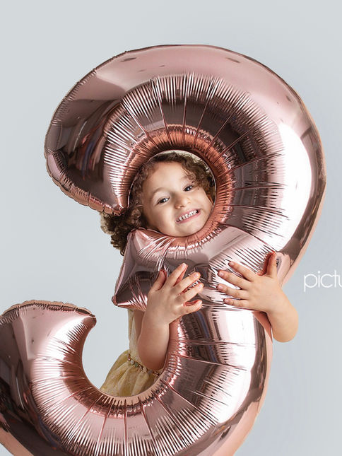 Smiling child peeking through a large rose gold number 3 balloon,Miami photo session Town gate