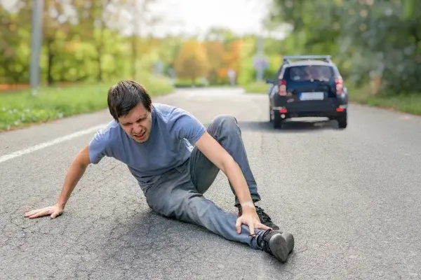 Man in gray clothes sits on a road, holding his leg in pain. A black car is in the background, with a green, blurry park setting.
