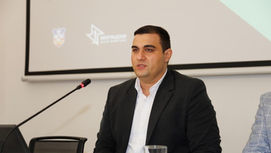 Miljan Ilić seated behind a conference desk during a business event, wearing a dark suit and white shirt while looking toward the audience.