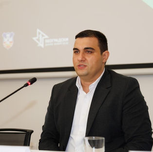 Miljan Ilić seated behind a conference desk during a business event, wearing a dark suit and white shirt while looking toward the audience.