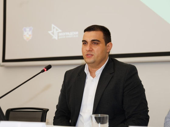 Miljan Ilić seated behind a conference desk during a business event, wearing a dark suit and white shirt while looking toward the audience.