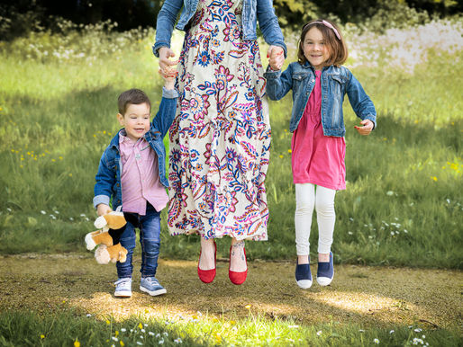 Une maman en robe fleurie saute avec ses deux jeunes enfants dans un champ verdoyant, lors d’une mini-séance photo à Chemillé-en-Anjou.
