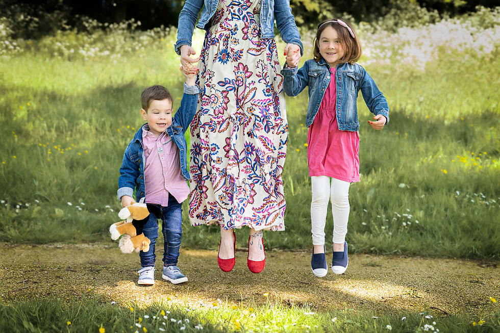 Une maman en robe fleurie saute avec ses deux jeunes enfants dans un champ verdoyant, lors d’une mini-séance photo à Chemillé-en-Anjou.