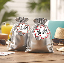 Two silver "Oh Sh*t Kit" drawstring bags on a wooden table with sunglasses and a drink.