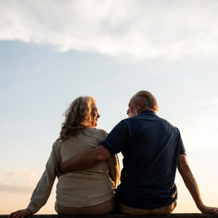 Elderly couple sitting on a ledge, embracing and gazing into each other's eyes during sunset. Calm, warm ambiance with cloudy sky.