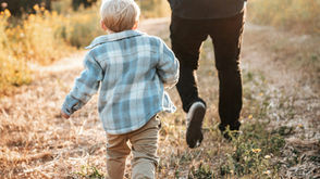 Child in a blue plaid shirt and adult running on a sunlit path surrounded by wildflowers, creating a joyful, serene scene.