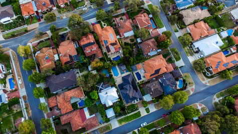Aerial view of a suburban neighbourhood with red-roofed houses, green trees, and winding roads. Pools are visible in some backyards.