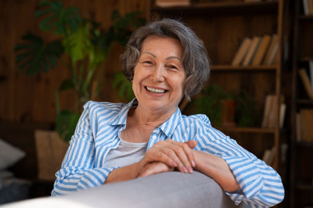 Smiling elderly woman in blue-striped shirt sits on couch, wooden bookshelf and plants in background, creating a cozy, serene setting.