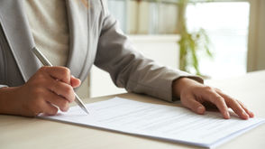 Person in a light gray suit writing on a document at a desk with a pen in hand. Bright office setting with blurred shelves and plant.