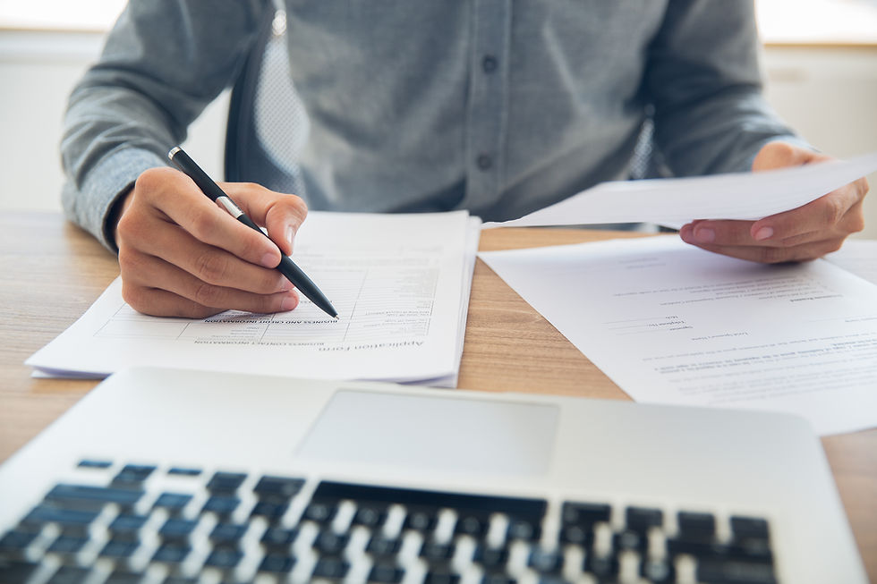 Person filling out forms at a desk with a pen in hand, holding papers. A laptop is visible in the foreground. Casual setting.