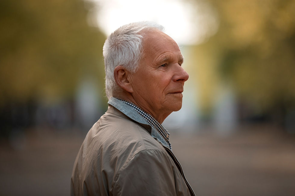 Elderly man in beige jacket gazes thoughtfully into distance. Soft focus background with warm, blurred greenery suggests a peaceful setting.