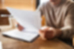 Person in a beige sweater reviewing documents at a wooden table, with blurred blinds in the background. Bright light creates a focused mood.