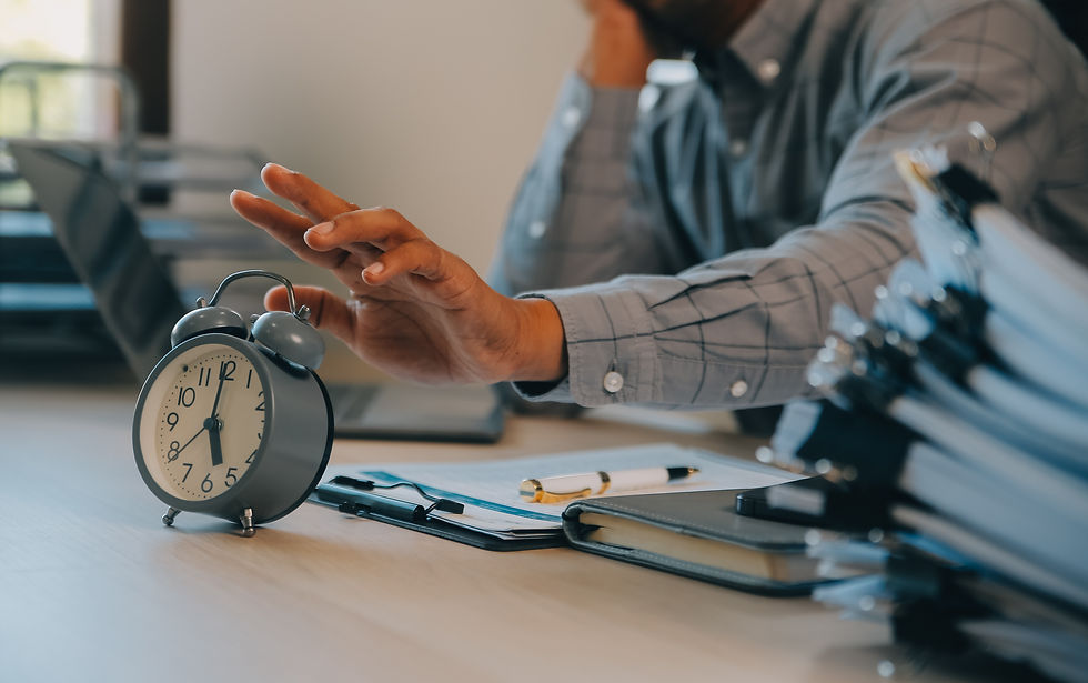 A person in a plaid shirt reaches to turn off a gray alarm clock on a desk with documents and a pen. The setting is an office.
