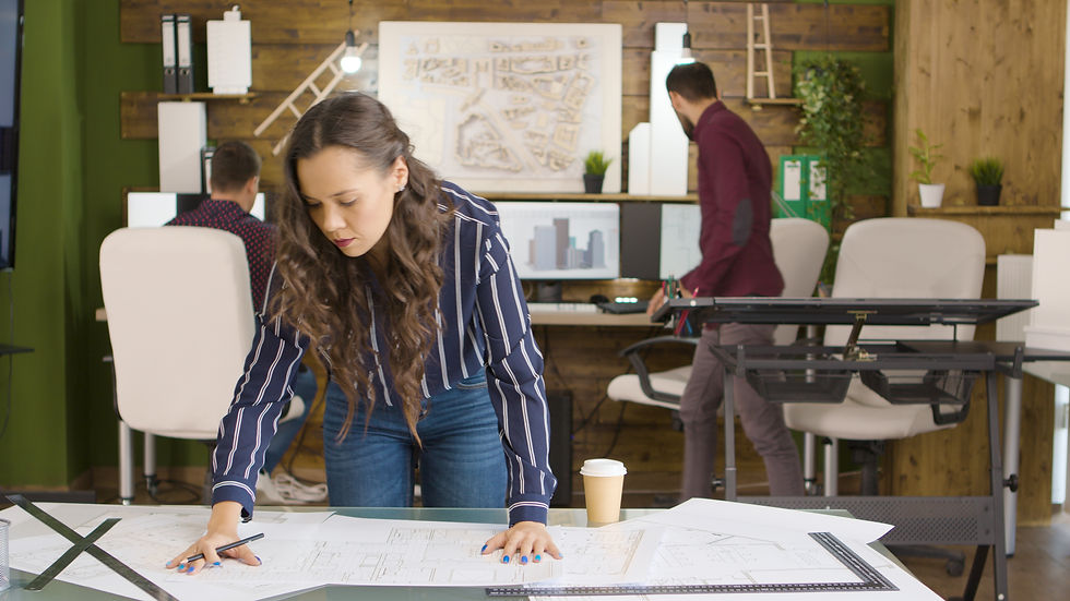 Woman reviewing architectural plans on a desk in a modern office, two colleagues in the background. Wood panel walls and green decor.