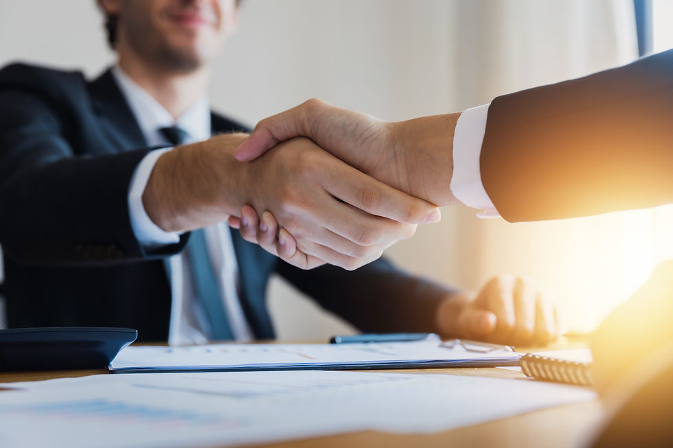 Two people shake hands over a desk with documents, pens, and a calculator. Sunlight streams in, creating a warm, professional setting.