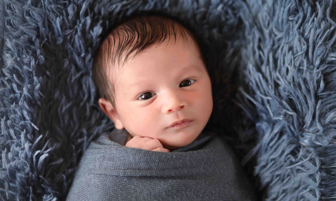 An alert, awake newborn baby with open eyes wrapped in a charcoal grey swaddle, resting on a plush blue fur backdrop during an in-home photo session in Atlanta.