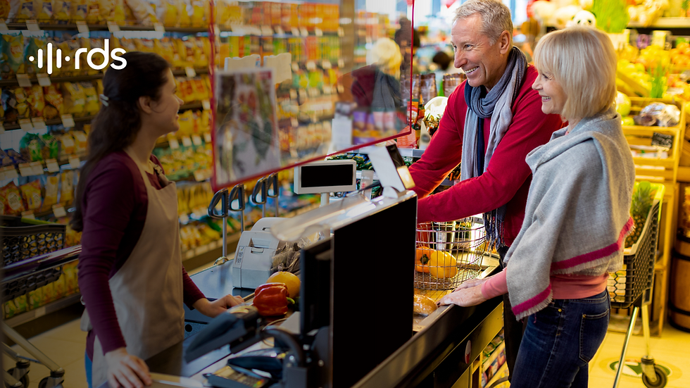 Casal sorridente interage com atendente em caixa de supermercado, durante o pagamento das compras, em um ambiente colorido e acolhedor, com destaque para a proximidade e simpatia no atendimento.
