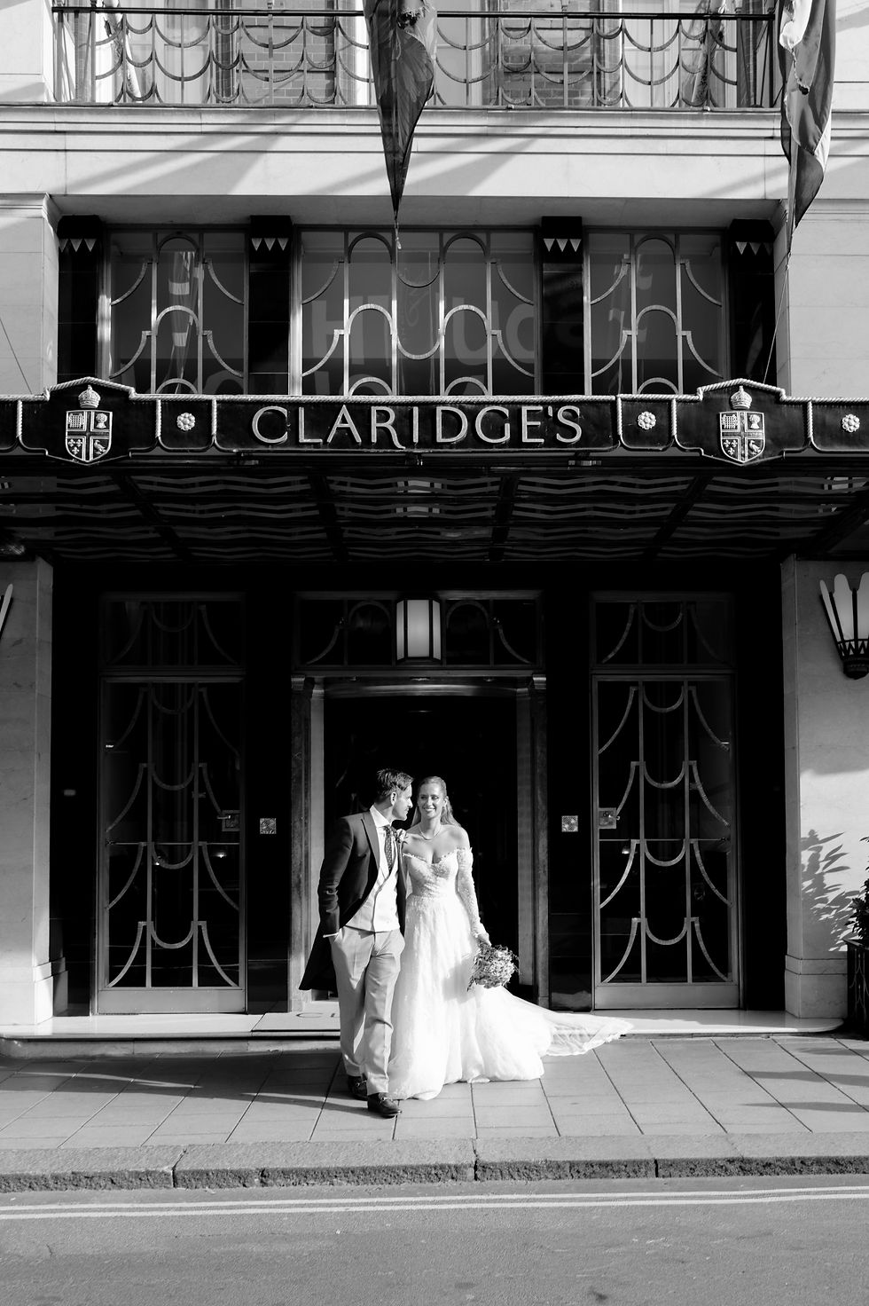 Bride and groom in formal attire smile outside Claridge's hotel. Black and white image with elegant facade and ornate details.