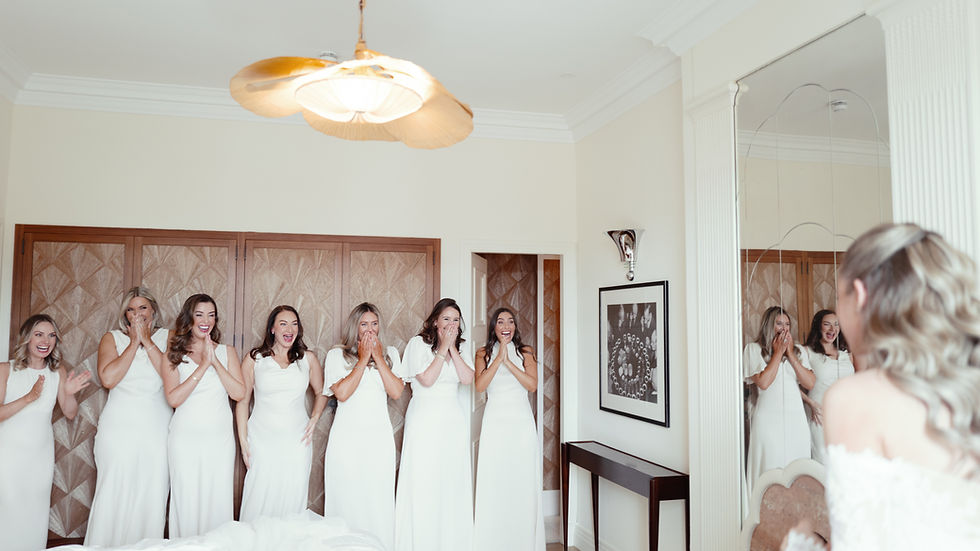 Bridesmaids in white dresses react excitedly as they look at the bride in a mirror. Room features wood panel doors and a ceiling fan.