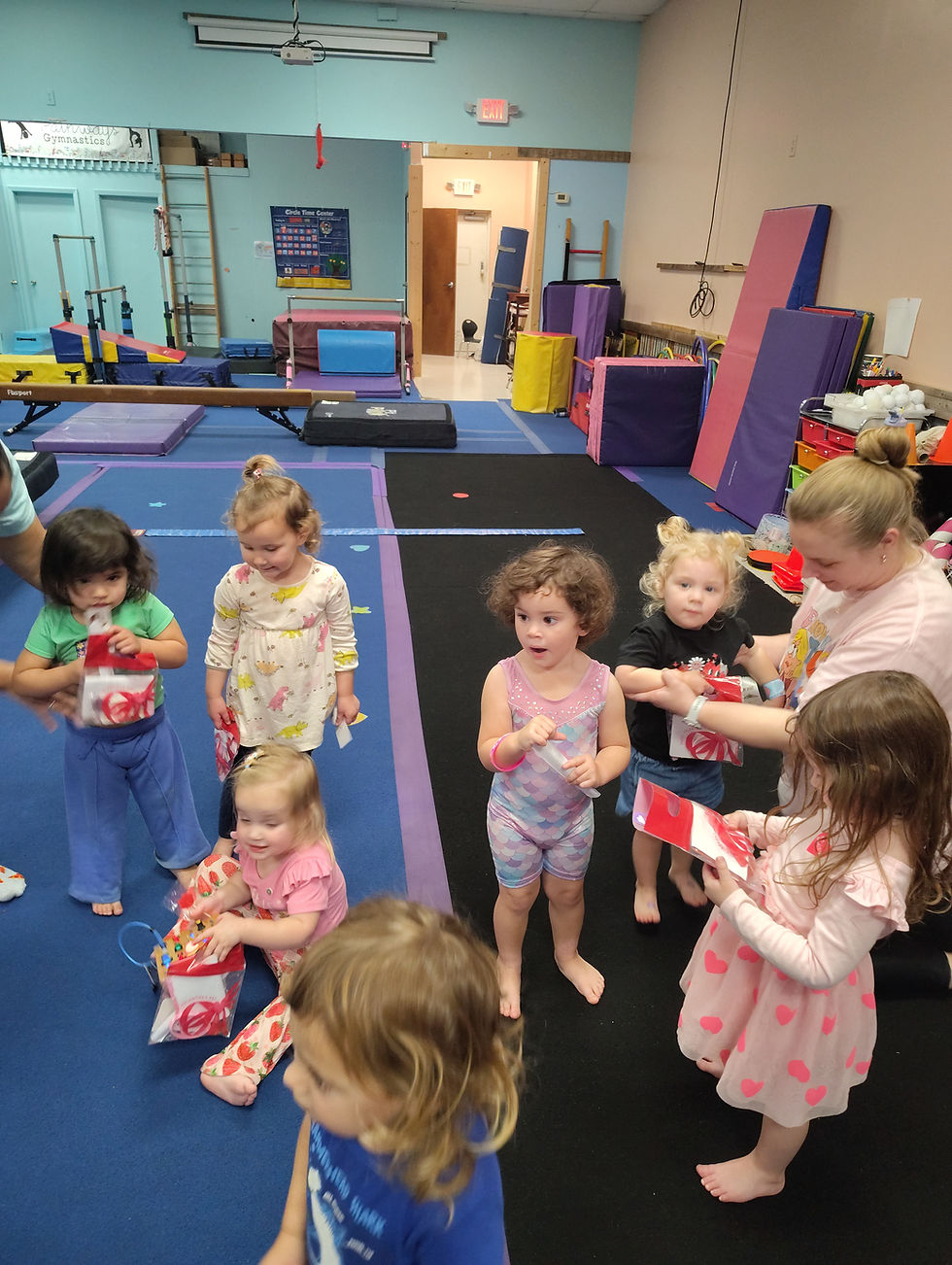 Children in a colorful gymnastics room hold red bags and chat. Mats and equipment are visible. The mood is playful and lively.